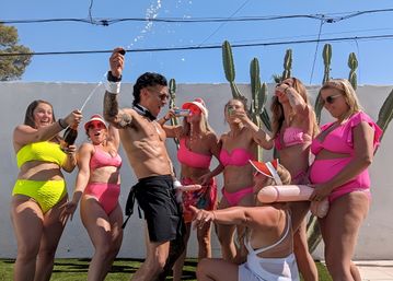 Shirtless person spraying champagne as a laughing group in bright bikinis drinks and poses with a novelty inflatable in a sunny cactus-lined backyard pool party
