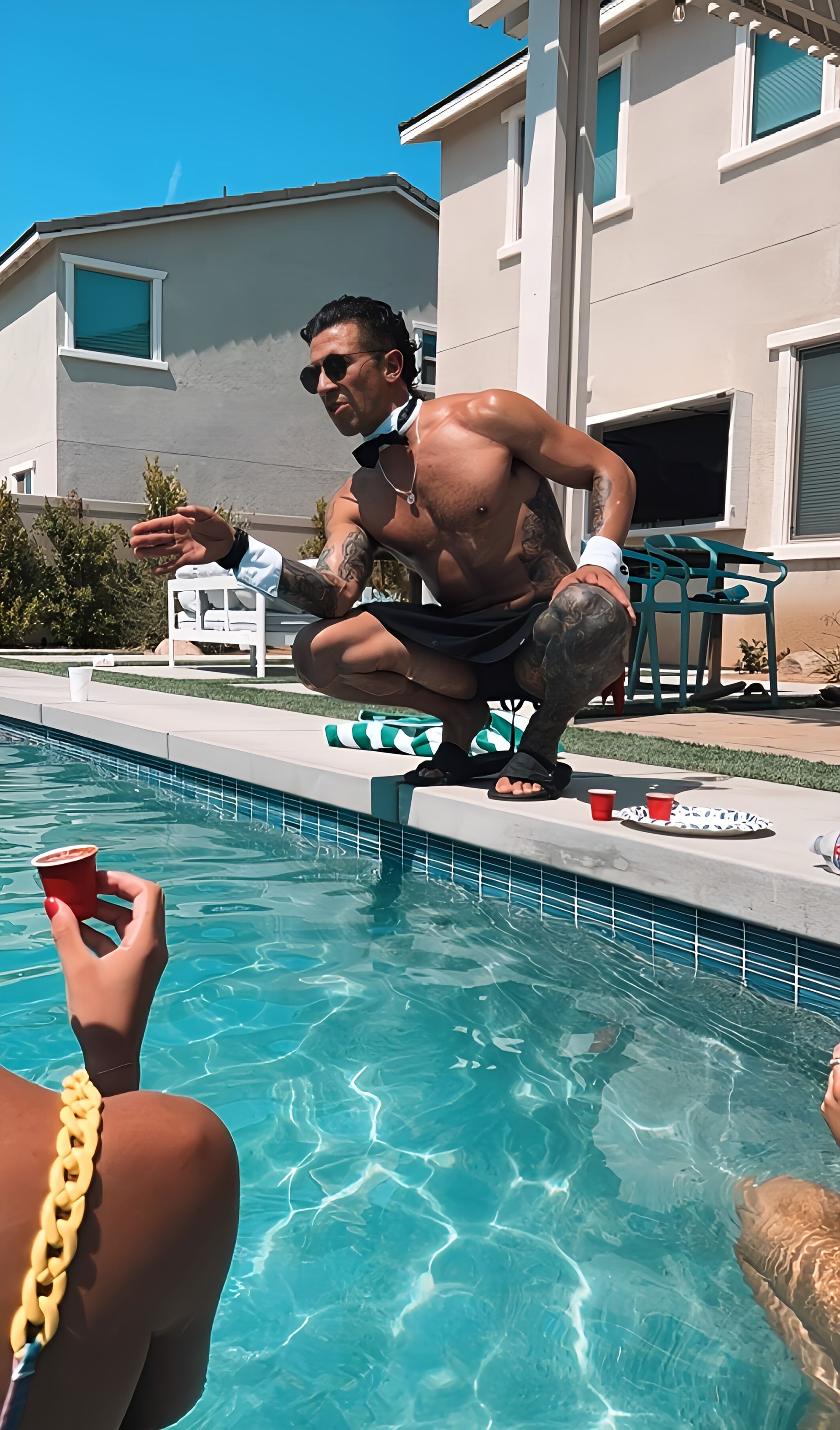 Shirtless entertainer in sunglasses, cuffs and bow tie crouches at the edge of a suburban backyard pool offering a red shot cup to a guest in the water on a sunny summer pool day.
