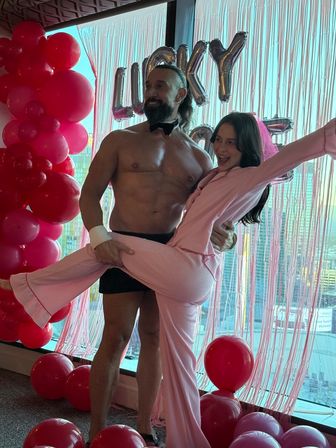 Playful bachelorette party in a hotel suite: shirtless male entertainer in bow tie lifts a smiling woman in pink pajamas and veil amid pink balloons and a city skyline backdrop.