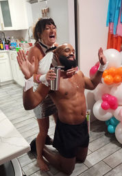 Playful indoor costume party in a kitchen: laughing woman in a white top and sequined skirt poses behind a kneeling shirtless man wearing cuffs and a collar, he holds a stainless tumbler, colorful balloon cluster and kitchen counters in the background.