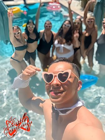 Sunlit outdoor pool party selfie of a person in heart-shaped sunglasses and a bow tie, with a cheering group in swimsuits and colorful floaties in the pool behind.