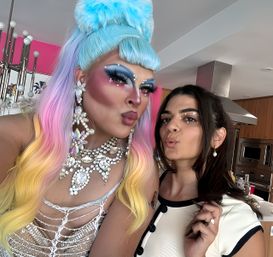 Selfie in a modern kitchen: a drag performer with a pastel rainbow wig, blue beehive, bold blue eye makeup and large crystal jewelry posing with a friend with dark hair, pearl earrings and subtle face gems, both making kissy faces.