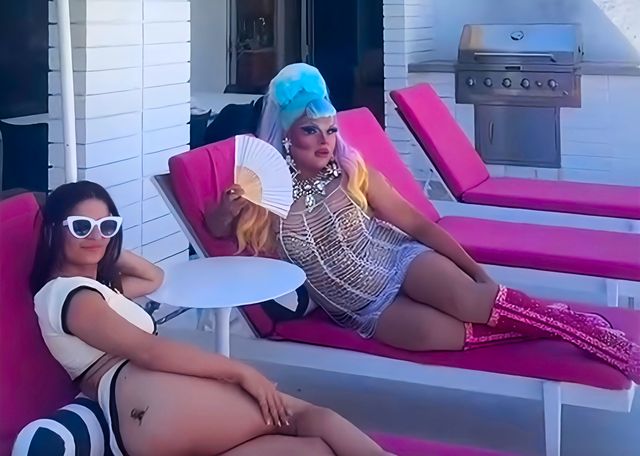 Poolside scene on a pink outdoor lounge: a person in white sunglasses and a swimsuit relaxes beside a glamorous drag performer in a blue wig, sequined dress, statement jewelry, pink glitter boots and a white hand fan, with a patio grill in the background.