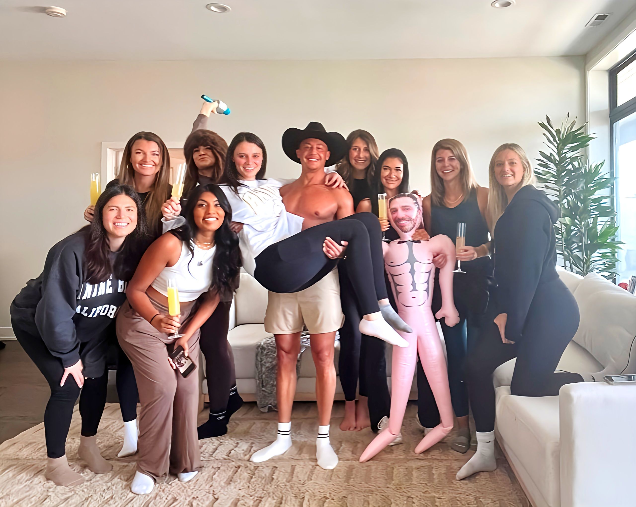 Group of friends in a bright apartment living room celebrating with mimosas — a shirtless man in a cowboy hat playfully lifts a woman while others smile and pose with an inflatable doll, bachelorette-style brunch vibe.
