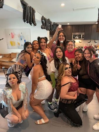 Fun group of friends in colorful costumes posing and making faces at an indoor costume party in a modern kitchen-living room with a black balloon banner and Halloween bat decorations.