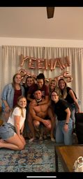 Smiling friends at an indoor bachelorette party in a living room, posing in front of rose-gold letter balloons and curtains with a woman perched on a shirtless man's shoulders for a playful group photo.