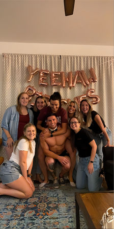 Friends group photo in a living-room party: eight women gathered around a shirtless man in a bow tie, smiling in front of rose-gold foil "YEEHAW" balloons and a patterned rug.