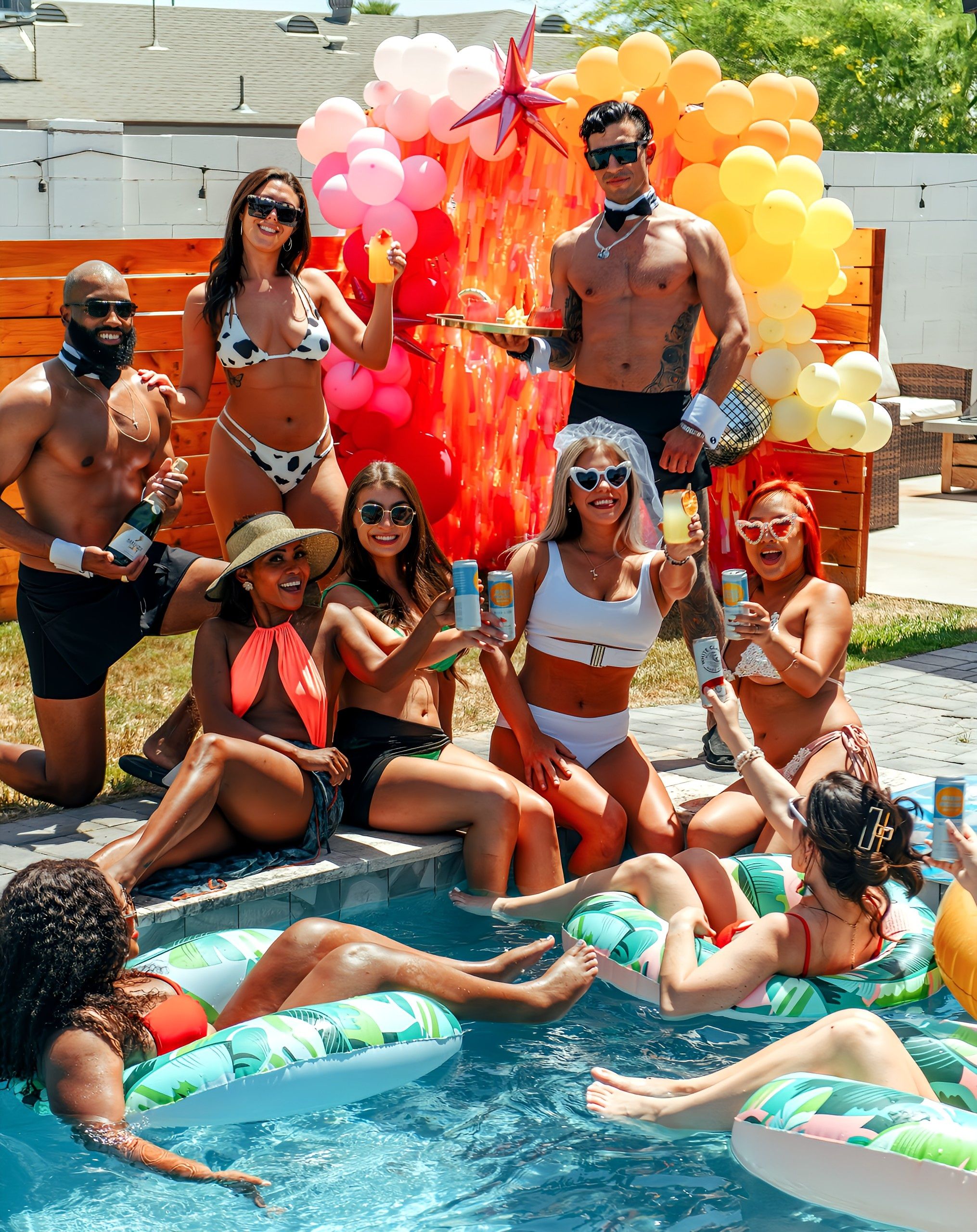 Sunny backyard summer pool party with friends in swimsuits toasting drinks by a pink-orange balloon arch, float tubes in the pool, one guest wearing a veil and heart sunglasses