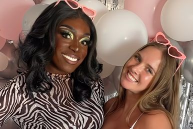 Two smiling friends pose at a party photo booth with pink and silver balloons; one in a zebra-print top and bold green eye makeup, the other in a white tank, both with pink heart sunglasses perched on their heads.