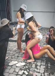 Women in bikinis and bucket hats at a lively backyard patio pool party, laughing and dancing while dollar bills and plastic cups scatter on wet pavers.