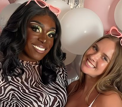Two smiling friends posing in a balloon photo booth with pink and white balloons, zebra-print top and pink heart-shaped sunglasses