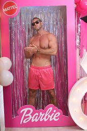 Shirtless muscular man in pink swim trunks and sunglasses posing inside oversized pink doll-box photo booth with silver tinsel backdrop, white balloons and marquee lights - playful party display