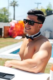 Smiling shirtless man in sunglasses and a black bow tie leaning at a sunny backyard poolside bar, colorful inflatables and a palm tree in the background at a summer pool party.