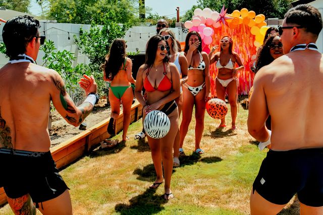 Sunny backyard summer party with friends in swimsuits and swim trunks laughing and playing with zebra and leopard-print beach balls in front of a pink-orange balloon arch and fringe backdrop on a grassy lawn.