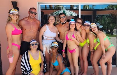 Pool-party group of friends in colorful swimsuits and visors posing and smiling outside a pink mid-century house with sliding glass doors and palm tree reflections