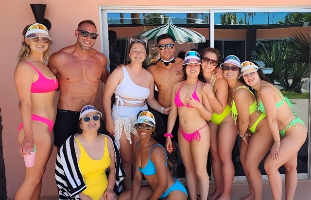 Smiling group of young adults in bright swimsuits and visors at a sunny poolside patio, posing in front of sliding glass doors with a striped umbrella reflection.