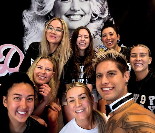 Eight smiling people take a close-up group selfie in front of a large black-and-white portrait mural inside a studio, casual friendly team vibe.