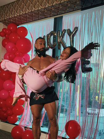 Muscular man in a bow tie and apron lifts a laughing woman in pink pajamas at a festive high-rise party with a tall pink balloon column, metallic letter balloons spelling "LUCKY", pink fringe backdrop and a city skyline visible through the window.