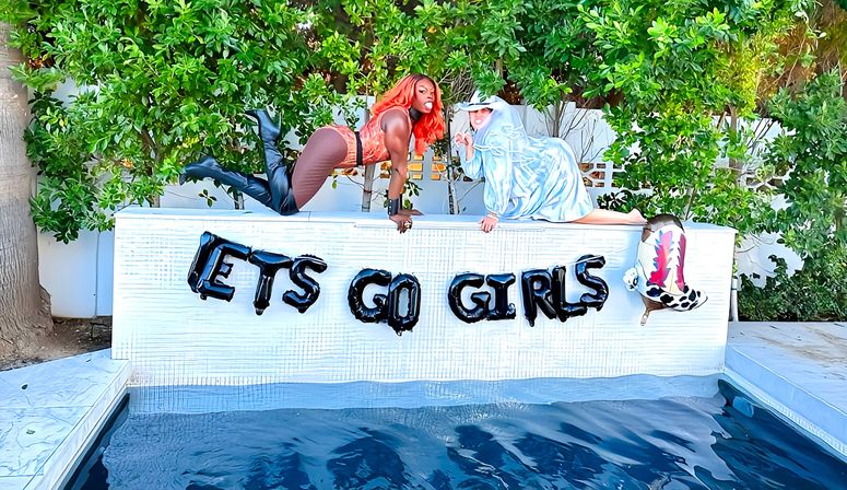 Backyard pool party scene with two people playfully posing on a white pool ledge above a blue pool, black letter balloons reading "ETS GO GIRLS", lush green hedges and a decorative cowboy boot balloon