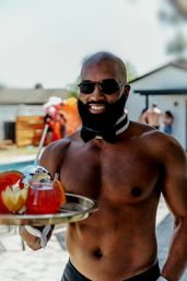 Poolside shirtless server wearing a bow tie and sunglasses holding a tray of colorful cocktails and carved fruit at a sunny backyard pool party