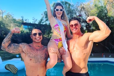 Backyard pool party on a sunny day — woman in a colorful bikini and sash lifted and cheered by two shirtless, sunglasses-wearing muscular men showing tattoos.