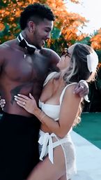 Romantic couple embracing by a backyard pool at sunset — a shirtless man with a bow tie and tattoos smiles at a woman in a white swimsuit and tied sarong with orange foliage glowing behind them.