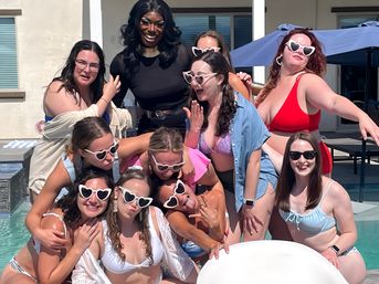 Sunny pool party scene: a lively group of friends in swimsuits and heart-shaped sunglasses laughing and posing together around a tall performer in black on a resort-style patio by the pool.