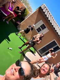 Fun group selfie of people in activewear doing an outdoor workout on bright green turf in a sunny suburban backyard by a tan stucco house, cheering and flashing peace signs.