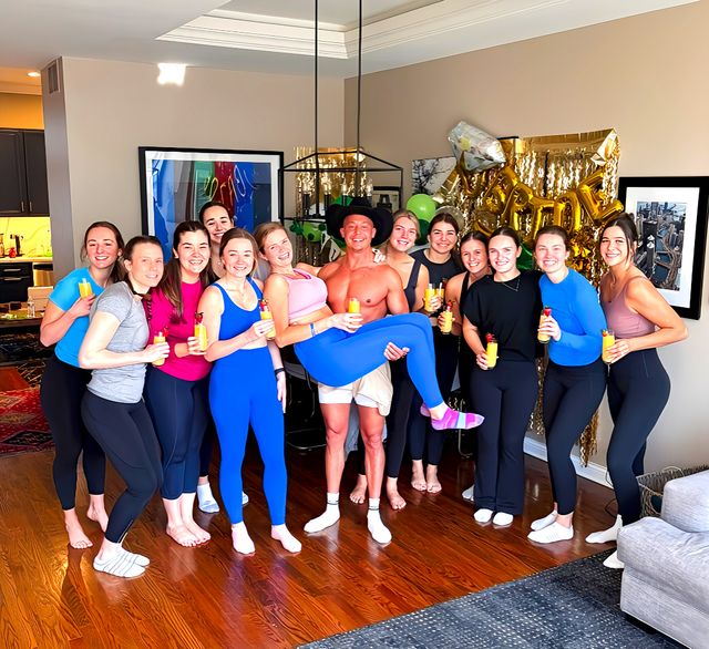 At-home bachelorette-style brunch in a living room: a group of friends in activewear smiling and holding mimosas, gold celebratory balloons in the background and a shirtless man playfully lifting a guest.