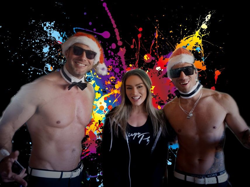 Smiling woman flanked by two shirtless men in Santa hats, sunglasses and bow-tie collars posed in front of a vibrant multicolor paint-splash backdrop — festive party vibe.