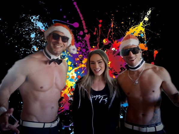 Smiling woman flanked by two shirtless men in Santa hats, sunglasses and bow-tie collars posed in front of a vibrant multicolor paint-splash backdrop — festive party vibe.