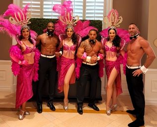 Six cabaret-style performers posing in a hotel ballroom: three women in bright pink sequined showgirl costumes with feathered headdresses and three shirtless male dancers in black trousers, white cuffs and bow ties.