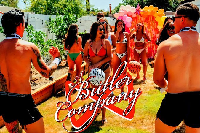 Sunny backyard summer party with bikini-clad guests and two shirtless men playing with beach balls in front of a colorful balloon arch