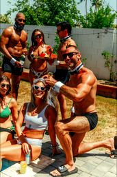 Sunny backyard pool party with a bachelorette in a white swimsuit and heart-shaped sunglasses laughing as shirtless servers in black shorts and bow ties offer cocktails and grapes poolside