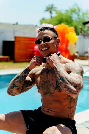 Muscular tattooed man in sunglasses adjusts a bow tie poolside at a backyard summer party with a colorful balloon arch and palm tree in the background