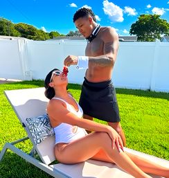 Sunlit suburban backyard scene: woman in a white swimsuit lounges on a chair while a shirtless man in a bow tie, cuffs and black apron feeds her a strawberry on the green lawn.