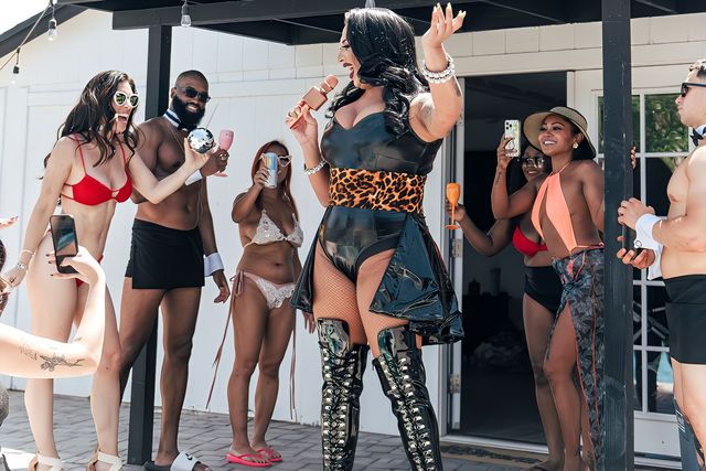 Drag performer in black vinyl and leopard waist belt singing with a microphone at a sunny backyard poolside party, surrounded by guests in swimsuits holding drinks and phones under a cabana.