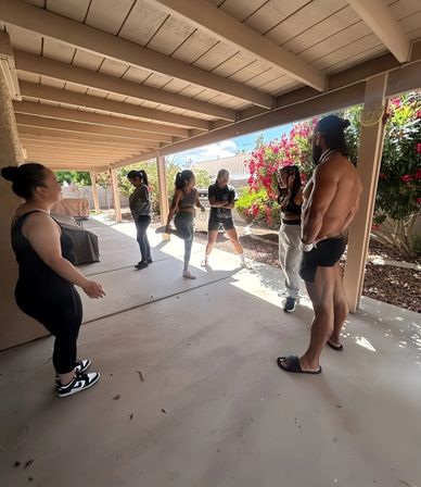 Group of six people chatting and stretching under a shaded backyard covered patio with wood beams, flowering shrubs and a sunny suburban yard.