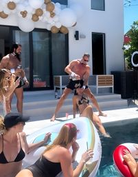 Sunny luxury backyard pool party at a modern white house with guests in swimsuits on inflatables, a shirtless performer pouring champagne over a seated woman, and white-and-gold balloon garland over the patio.