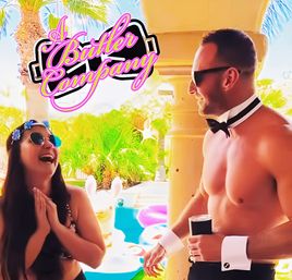 Sunny pool party at a tropical resort — laughing woman in sunglasses and flower crown greets a shirtless pool attendant in bow tie and cuffs holding a drink, palm trees and colorful inflatables nearby, retro pink script logo overhead.