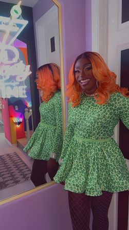 Smiling person with vibrant orange hair wearing a green leopard-print mini dress and fishnet tights, posing beside a gold-framed mirror in a colorful boutique dressing room with neon decor
