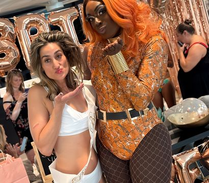 Two guests pose at an indoor rose-gold bachelorette party — woman in a white sash and swimwear blowing a kiss beside a performer in an orange wig, metallic patterned bodysuit, fishnet tights and gold cuffs, with rose-gold balloons and sequin backdrop.