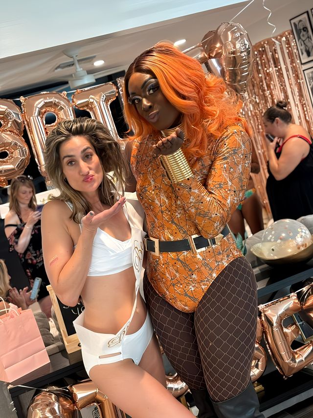 Two guests pose at an indoor rose-gold bachelorette party — woman in a white sash and swimwear blowing a kiss beside a performer in an orange wig, metallic patterned bodysuit, fishnet tights and gold cuffs, with rose-gold balloons and sequin backdrop.