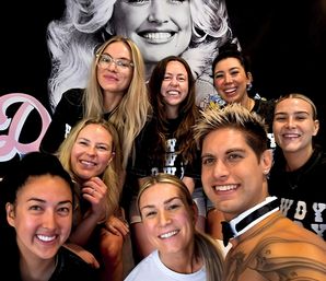 Close-up group selfie of eight smiling adults posing indoors in front of a large black-and-white portrait backdrop, cheerful crew vibe.