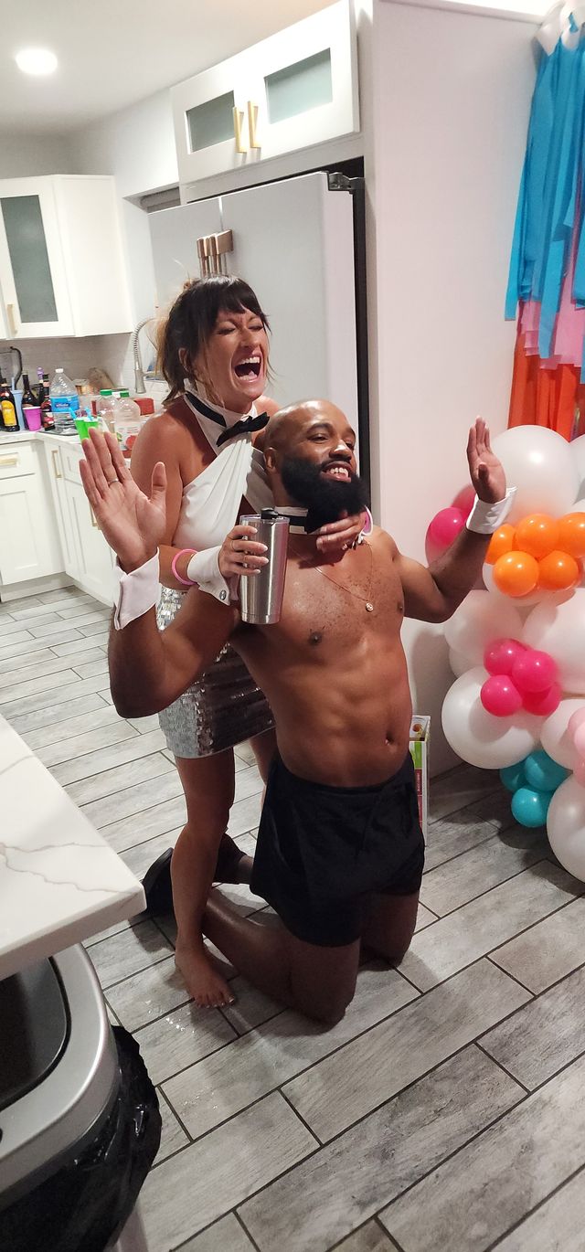 Playful indoor kitchen party: laughing woman in white top and sequined skirt playfully grabbing a kneeling shirtless man holding a tumbler, colorful balloons and kitchen cabinets in the background.
