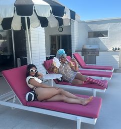 Two people lounging on bright pink chaise lounges at a sunny modern poolside patio — one in a bikini with white cat-eye sunglasses, the other in a blue wig, sequin dress, pink glitter boots and a hand fan under a black-and-white striped umbrella.