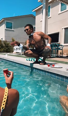 Shirtless tattooed man in a bow tie, cuffs and sunglasses crouching at a suburban backyard pool reaching toward swimmers holding red shot cups on a sunny summer day.