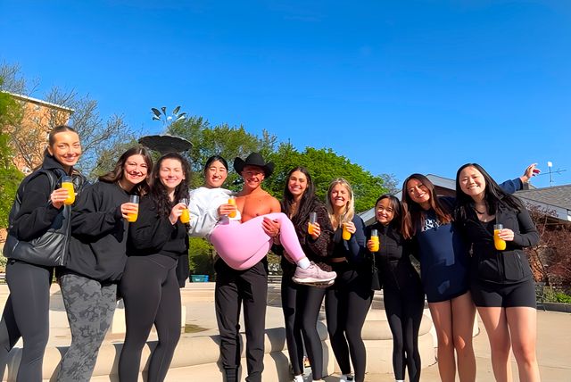 Group of young adults posing by a college campus fountain under a bright blue sky, holding orange drinks while a cowboy-hatted person lifts a friend in pink leggings.