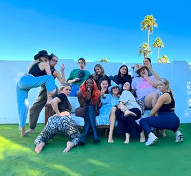 Playful group of friends posing on an outdoor sofa on a green lawn under a bright blue sky with palm trees, wearing casual clothes, cowboy hats and boots, central figure with bright red hair making a silly face
