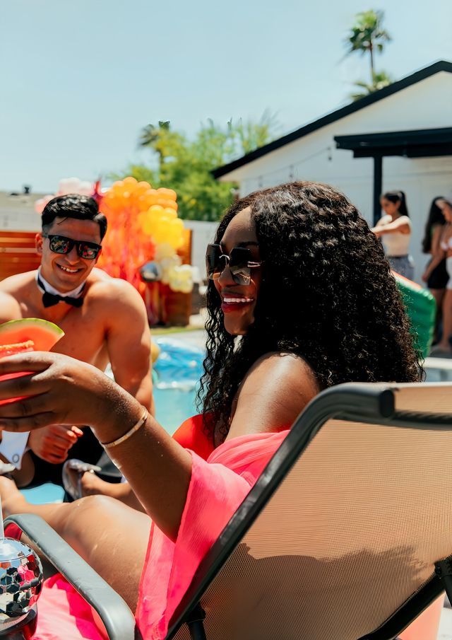Smiling woman in sunglasses and a bright pink cover-up holds a slice of watermelon poolside at a sunny backyard pool party, with a shirtless man in sunglasses and a bow tie and colorful balloons in the background.